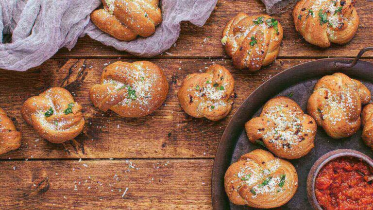 Garlic knots sprinkled with herbs and grated cheese are displayed on a wooden surface, alongside a bowl of tomato sauce—perfect movie night snacks for your next film marathon.