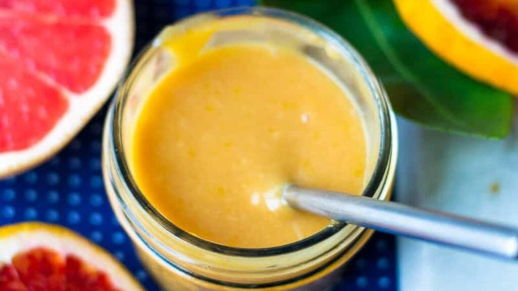 A glass jar filled with orange-colored creamy sauce or dressing, with a metal spoon inside. Sliced grapefruits are visible nearby on a blue surface.