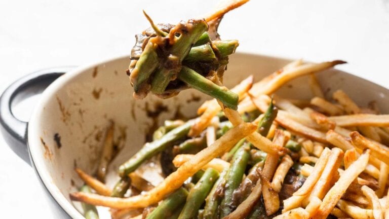 A casserole dish filled with cooked green beans and thin, crispy French fries, with a spoon lifting a portion of the mixture. Some fries and beans are coated in a dark sauce.