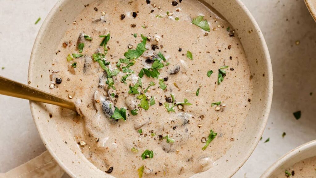 A bowl of creamy mushroom soup garnished with chopped parsley and cracked black pepper, with a spoon resting inside.