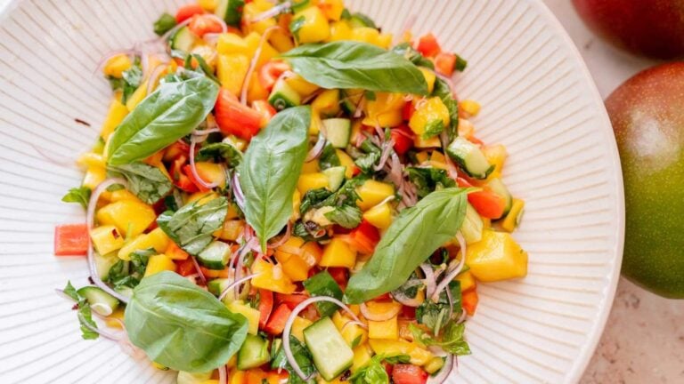A plate of mango salad with diced mango, cucumber, red bell pepper, sliced red onion, fresh basil leaves, and herbs on a white dish.