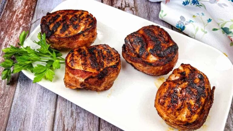 Four bacon-wrapped chicken breasts on a white rectangular plate garnished with sprigs of parsley.