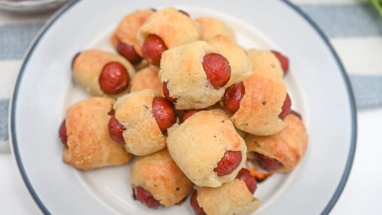 A plate of pigs in a blanket, featuring mini sausages wrapped in golden-brown pastry, sits on a white plate with a blue rim.