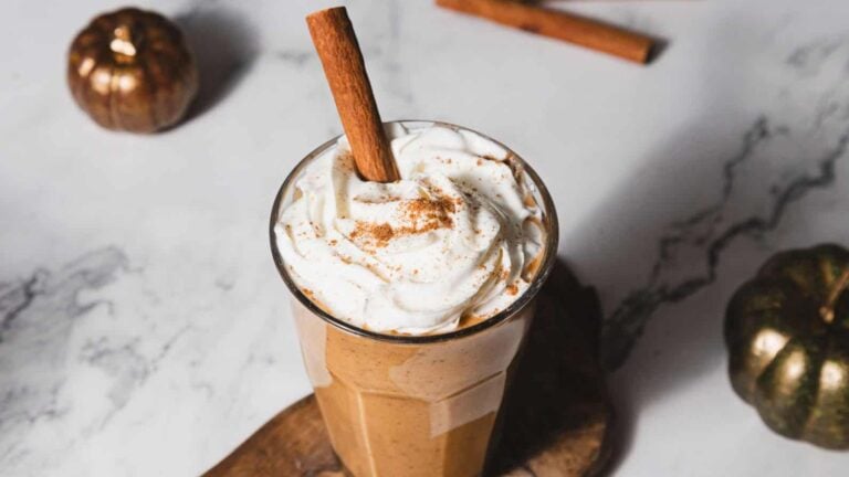 A tall glass filled with a creamy pumpkin spice latte topped with whipped cream and a cinnamon stick. The drink is set on a wooden coaster, and small decorative pumpkins are visible in the background on a marble surface.