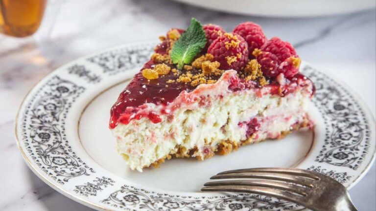A slice of raspberry cheesecake with a crumb crust, topped with fresh raspberries, mint, and crumbs, served on a decorative plate with a fork.
