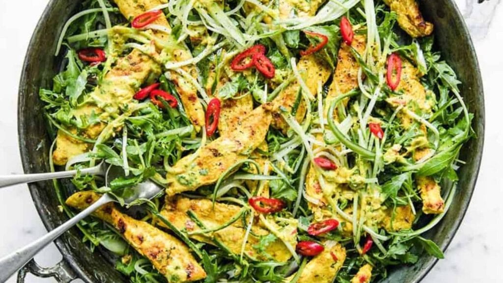 A skillet filled with grilled chicken strips, arugula, sliced red chili, and green herbs, served with metal tongs.