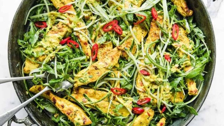 A skillet filled with grilled chicken strips, arugula, sliced red chili, and green herbs, served with metal tongs.