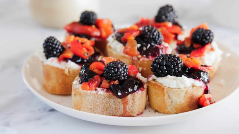 A plate of sliced baguette topped with ricotta cheese, blackberry jam, diced strawberries, and fresh blackberries, arranged on a white surface.