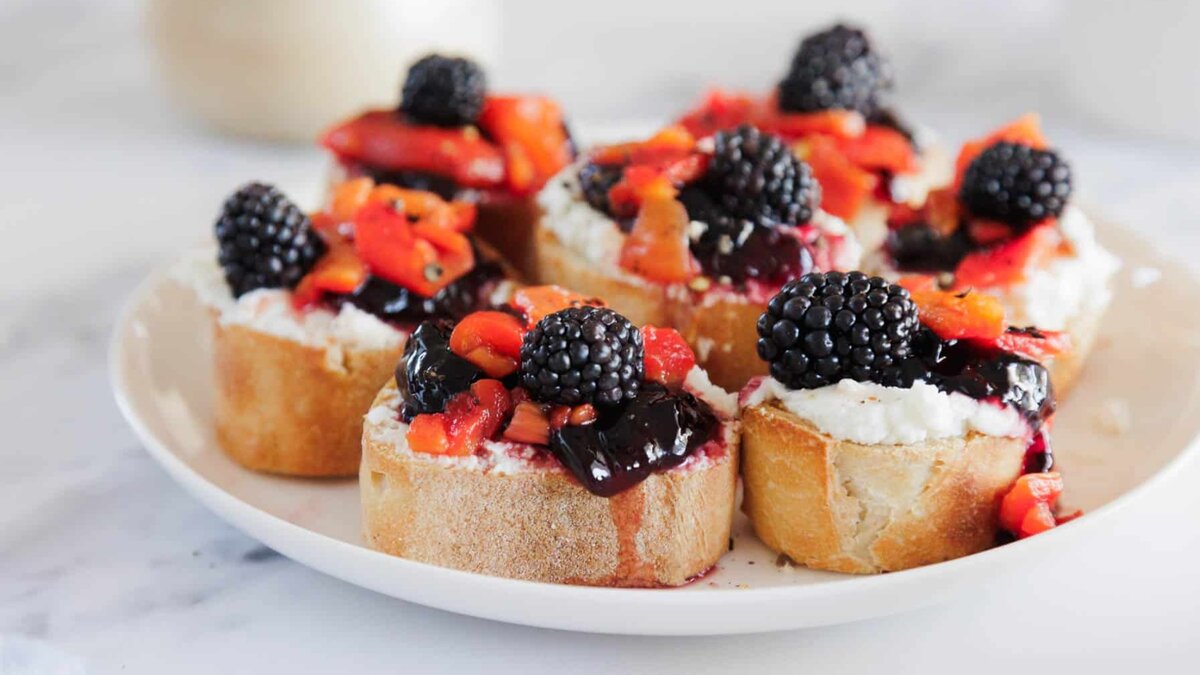 A plate of sliced baguette topped with ricotta cheese, blackberry jam, diced strawberries, and fresh blackberries, arranged on a white surface.