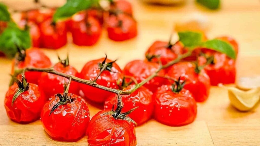 A cluster of roasted cherry tomatoes on the vine rests on a wooden surface, with a few green leaves and garlic cloves in the background.