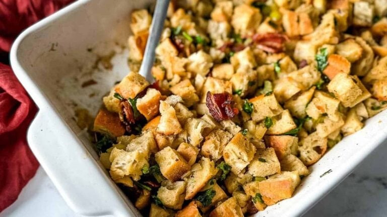 A white baking dish filled with cubed bread stuffing mixed with herbs and pecans, with a serving spoon inside.
