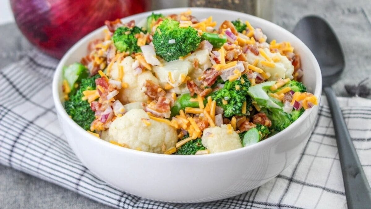 A bowl of broccoli cauliflower salad with shredded cheese and bacon, placed on a checkered cloth.