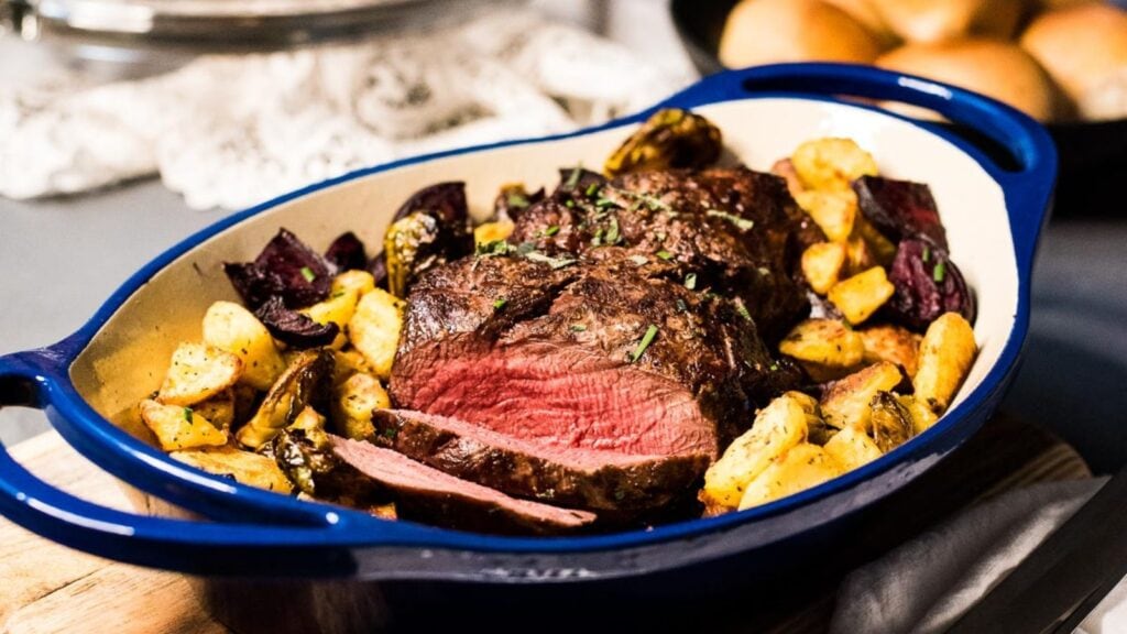 A blue baking dish with sliced roasted beef, golden potatoes, and vegetables, garnished with herbs, sits on a wooden board.