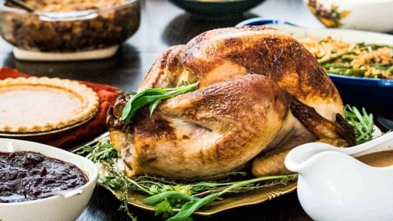 Roast turkey on a platter with herbs, surrounded by dishes of cranberry sauce, pie, green beans, and stuffing on a dining table.