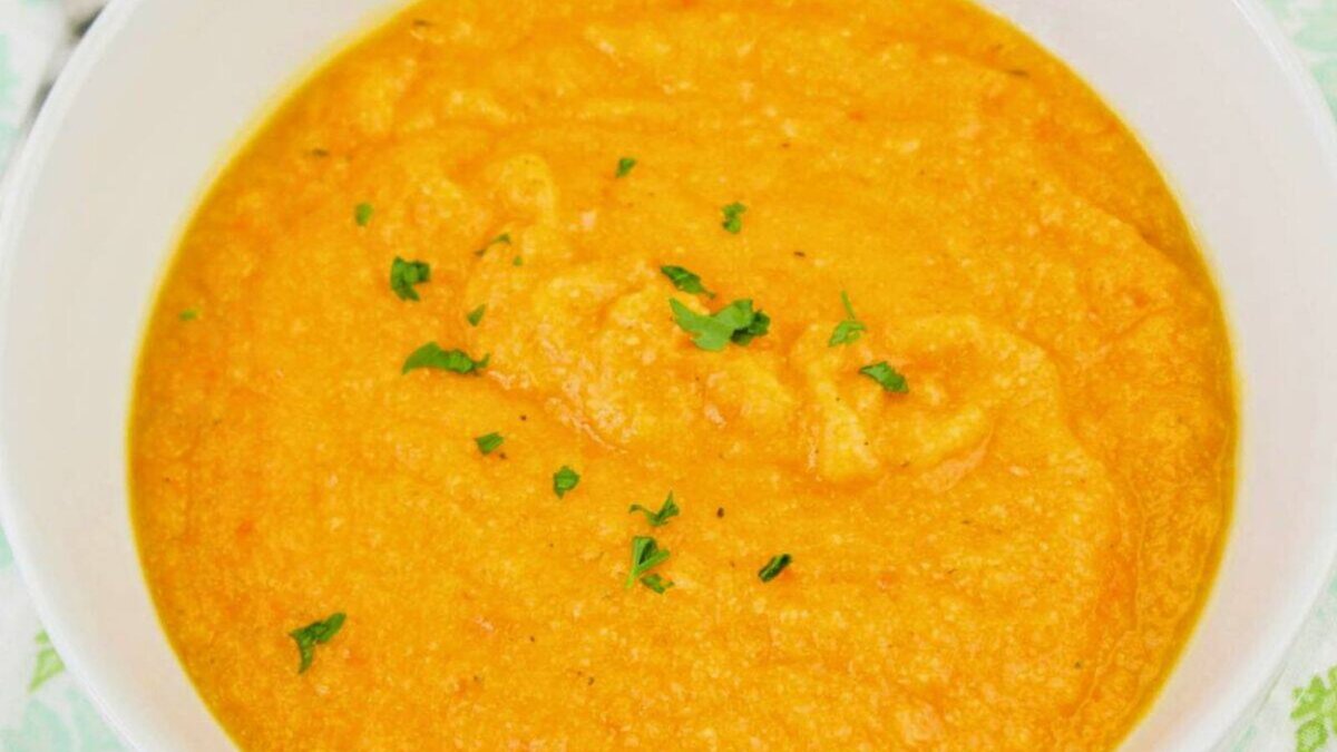 Bowl of creamy carrot soup garnished with small parsley pieces, placed on a patterned green and white napkin.
