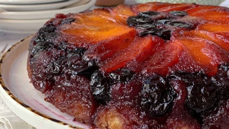 A close-up of a plum upside-down cake with caramelized plums and a glossy, dark fruit topping on a white plate.