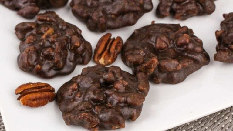 Five clusters of chocolate pecan pralines are arranged on a white plate, with two pecan halves placed nearby.