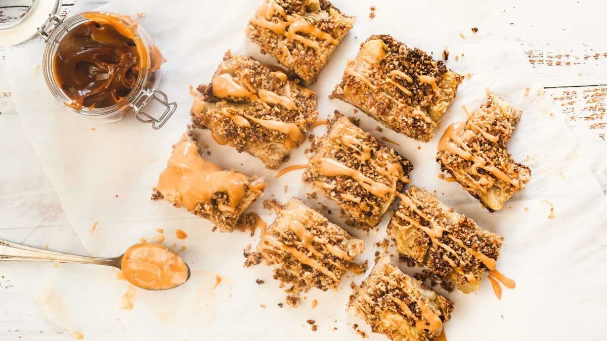 A batch of homemade apple crisp bars topped with a generous drizzle of caramel sauce, displayed on parchment paper with a jar of caramel and a spoon nearby.