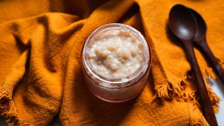 A glass jar filled with creamy rice pudding sits on an orange textured cloth next to two wooden spoons.