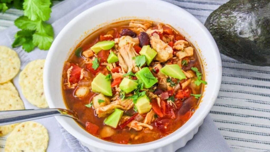 A bowl of chicken tortilla soup with diced avocado, beans, tomatoes, and herbs, beside tortilla chips and an avocado on a striped cloth.