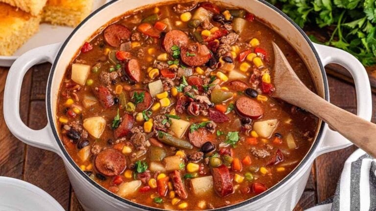 A pot of hearty soup with sausage, potatoes, corn, beans, and vegetables. A wooden spoon rests in the soup. Cornbread and parsley are visible in the background.