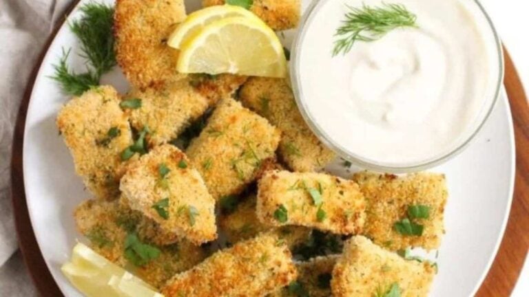 A plate of breaded fish sticks garnished with parsley and lemon wedges, served with a bowl of creamy white dipping sauce topped with dill.