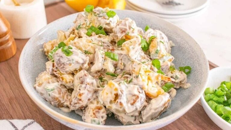 A bowl of creamy potato salad garnished with chopped green onions on a wooden table.