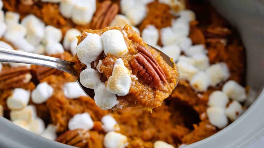 A spoonful of sweet potato casserole topped with mini marshmallows and a pecan, held over a dish filled with the same casserole.