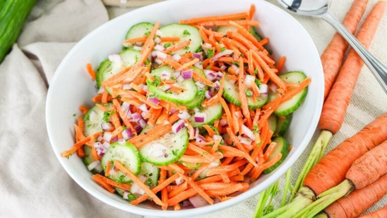 A white bowl filled with a salad of sliced cucumbers, shredded carrots, and chopped red onions, placed next to whole carrots on a cloth.