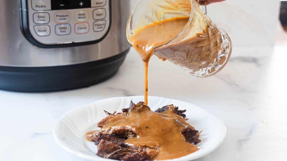 A person pours brown gravy from a glass container over shredded beef on a white plate. An electric pressure cooker is visible in the background on a white countertop.