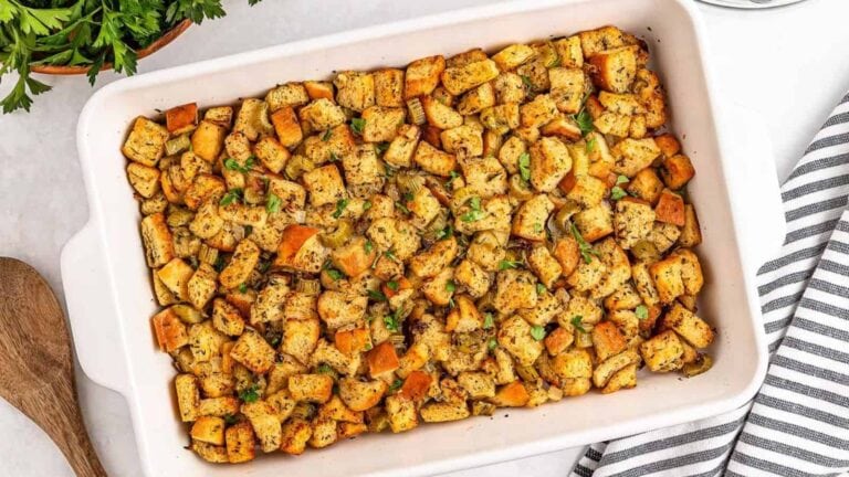 A rectangular dish filled with baked stuffing made of cubed bread and herbs, placed on a white surface next to a wooden spoon and a striped cloth.