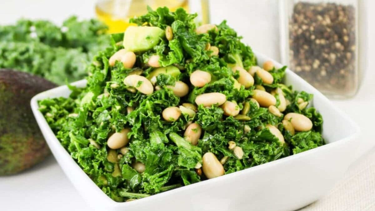 A square white bowl filled with kale salad, featuring beans and avocado pieces, placed on a table with a spoon beside it.