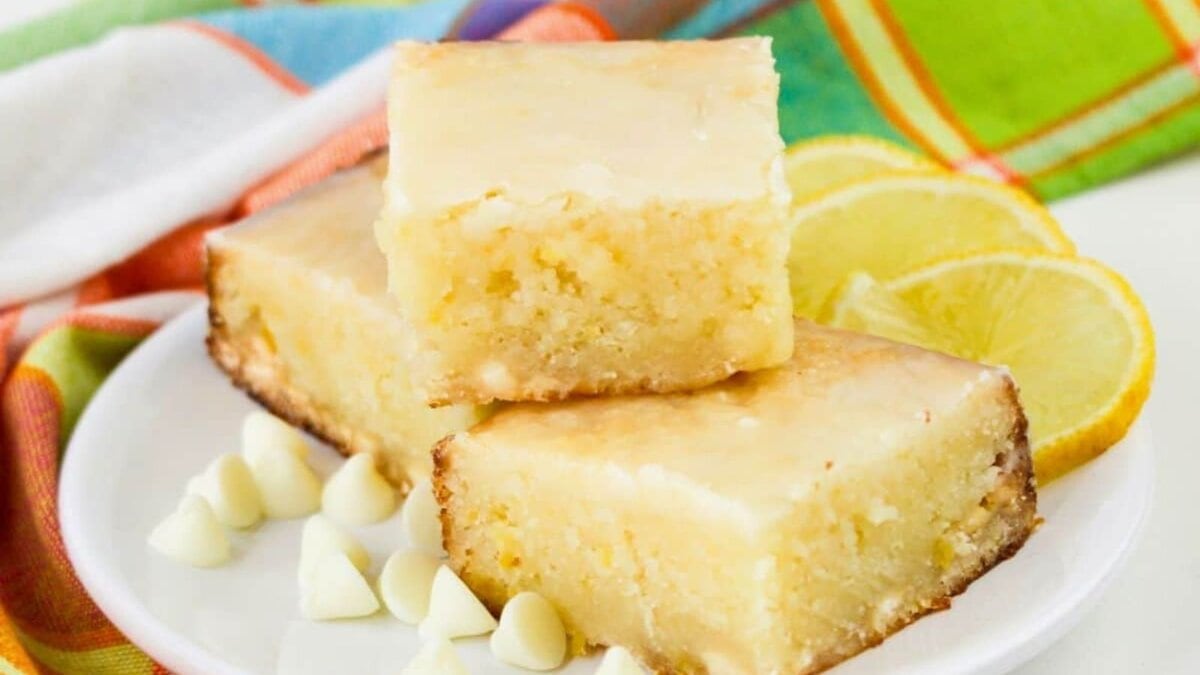 Three lemon bars stacked on a white plate, surrounded by white chocolate chips and lemon slices, with a colorful cloth in the background.
