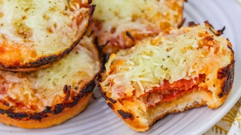 A close-up of mini pizza bagels topped with melted cheese, with one bagel bitten to reveal pepperoni and sauce inside, all arranged on a white plate.