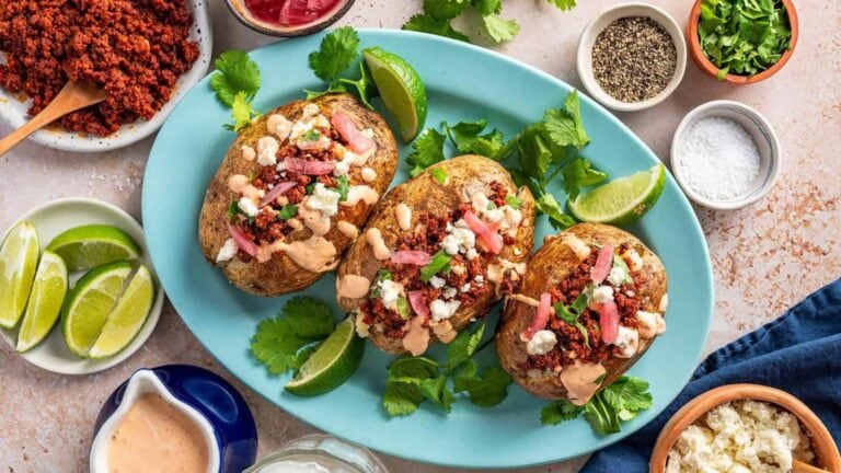 Three loaded baked potatoes topped with crumbled meat, cheese, and sauce on a blue platter, garnished with cilantro and lime wedges.