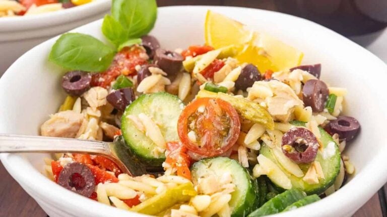 A bowl of pasta salad with sliced cucumber, tomatoes, olives, green beans, shredded chicken, and a lemon wedge, garnished with fresh basil.