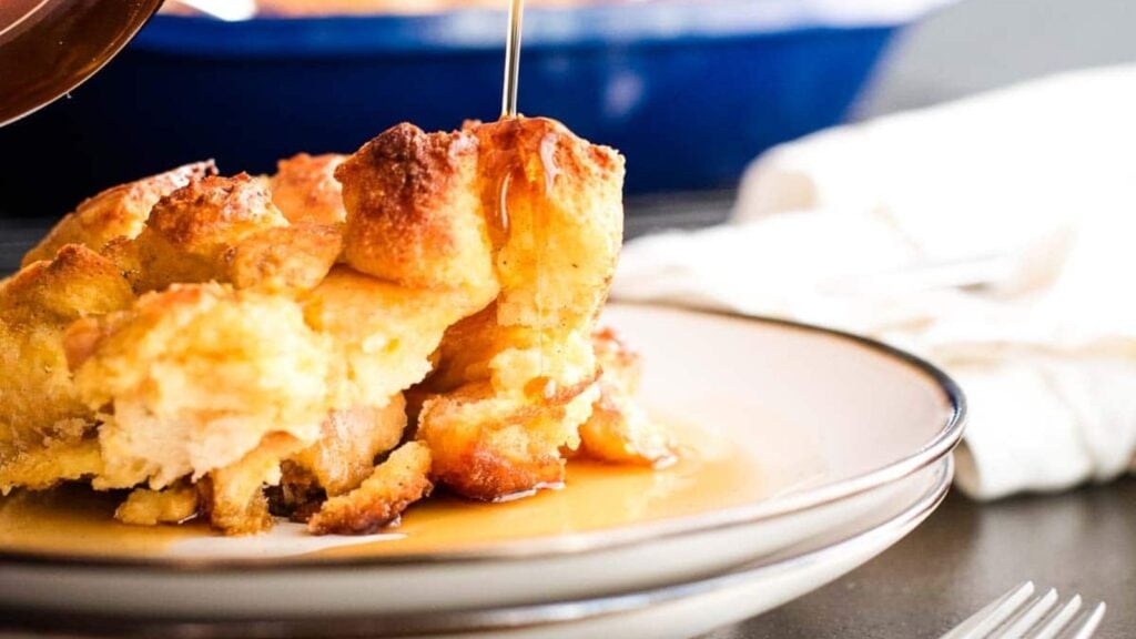 A close-up of bread pudding on a white plate with syrup being poured over it, with a fork and napkin in the background.