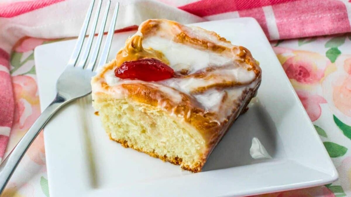 A slice of glazed peanut butter and jelly roll with a cherry on top sits on a white plate next to a fork, on a floral tablecloth.