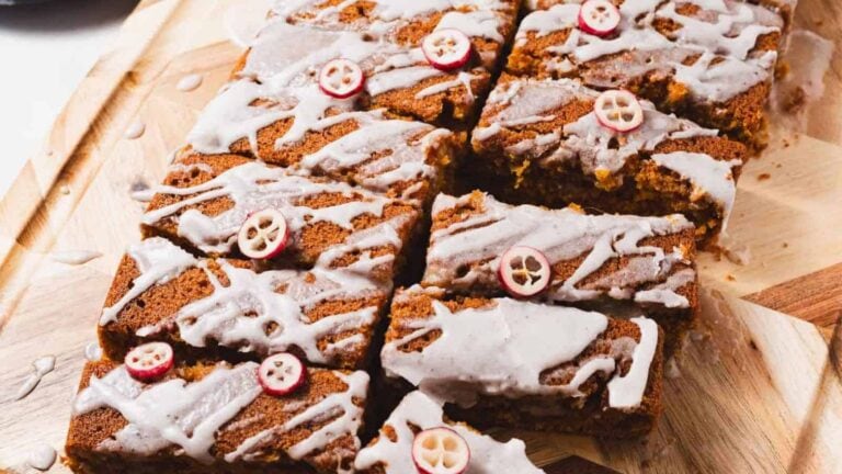 Sliced pumpkin bread topped with white icing and small cranberry slices, arranged on a wooden cutting board. The bread appears moist with a sprinkling of spices on top.