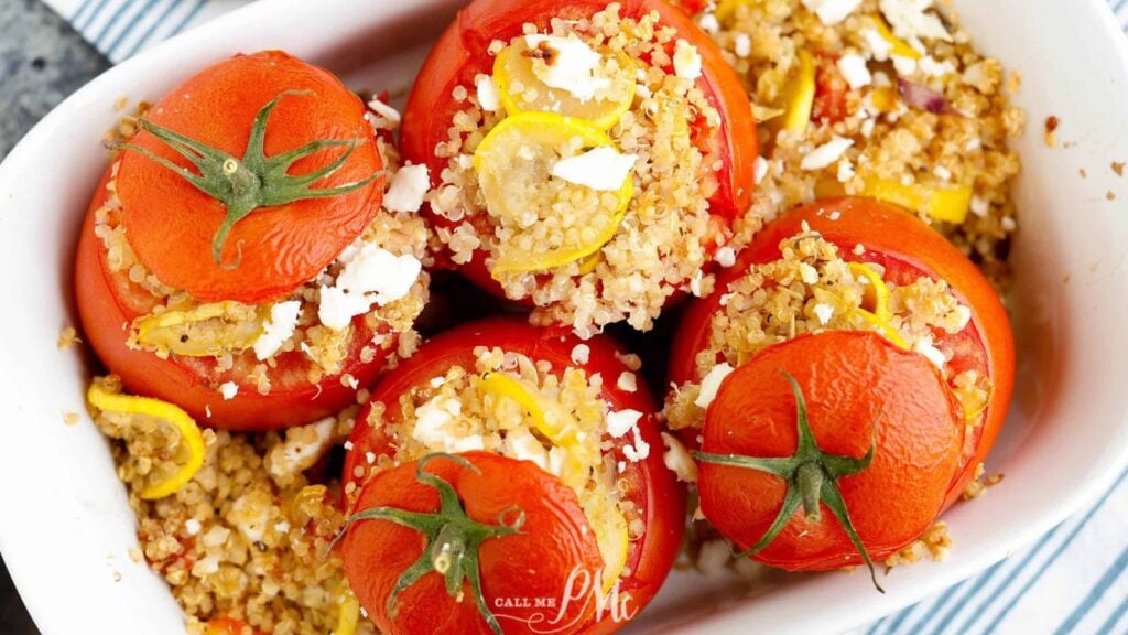 Five tomatoes stuffed with quinoa, yellow squash, and crumbled feta cheese are arranged in a white baking dish.