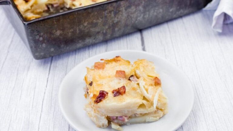 A serving of potato gratin with bacon is placed on a white plate, with the remaining casserole in a baking dish in the background on a light wooden surface.