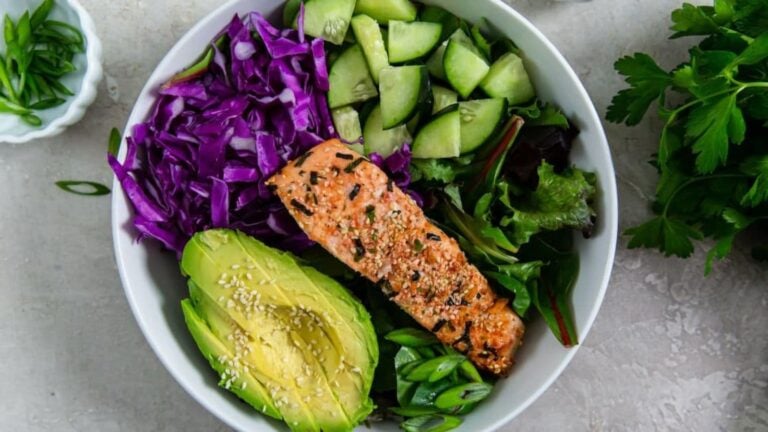 A bowl with sliced avocado, seared salmon, chopped cucumber, shredded purple cabbage, mixed greens, and green onions, garnished with sesame seeds.