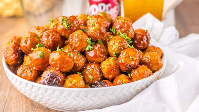 A white bowl filled with glazed meatballs garnished with chopped parsley, placed on a wooden table with a drink in the background.