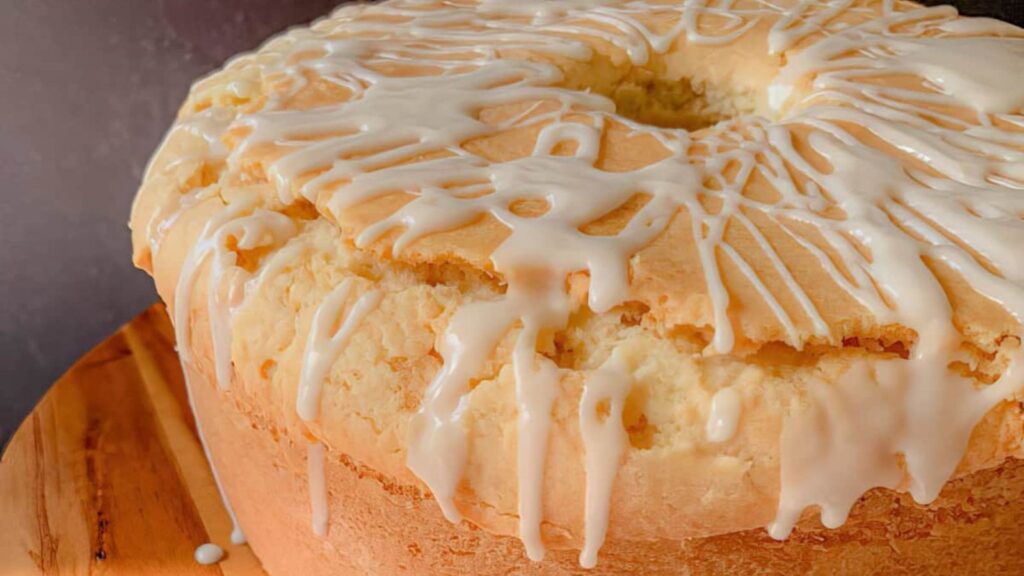 Round cake with white icing drizzled on top, sitting on a wooden surface. Cake has a cracked texture on the sides and a hole in the center.