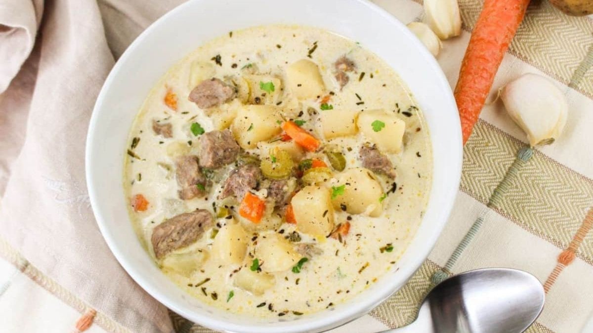 A bowl of creamy soup with chunks of meat, potatoes, and carrots, garnished with herbs. A spoon, carrot, garlic, and cloth napkin are nearby on a plaid tablecloth.