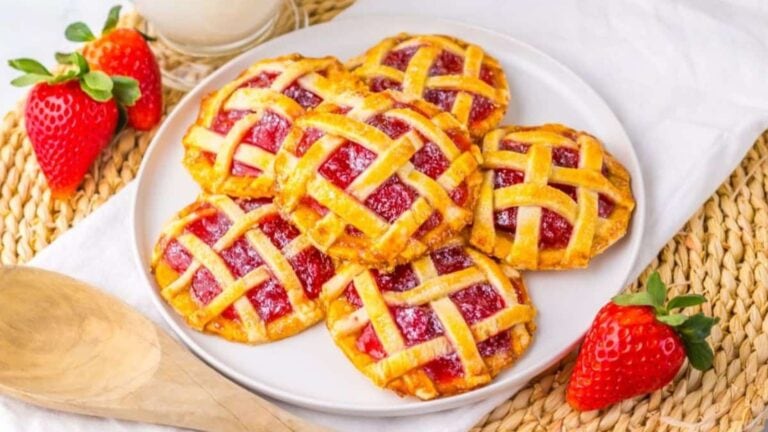A plate of lattice-topped strawberry pastries on a woven mat with fresh strawberries and a wooden spoon nearby. A glass of milk is in the background.