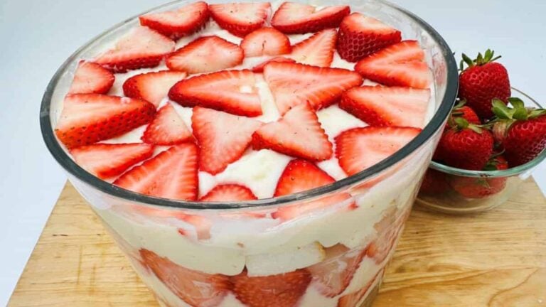 A glass bowl filled with layered strawberry trifle, topped with sliced fresh strawberries, sits on a wooden surface next to a small bowl of whole strawberries.