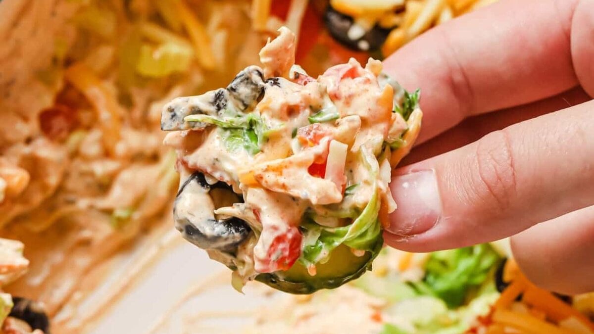 A hand holding a slice of cucumber topped with taco dip with cream cheese.