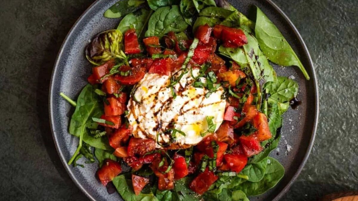 An assembled tomato burrata salad with sliced basil and burrata on a cutting board and a bowl of chopped heirloom tomatoes.