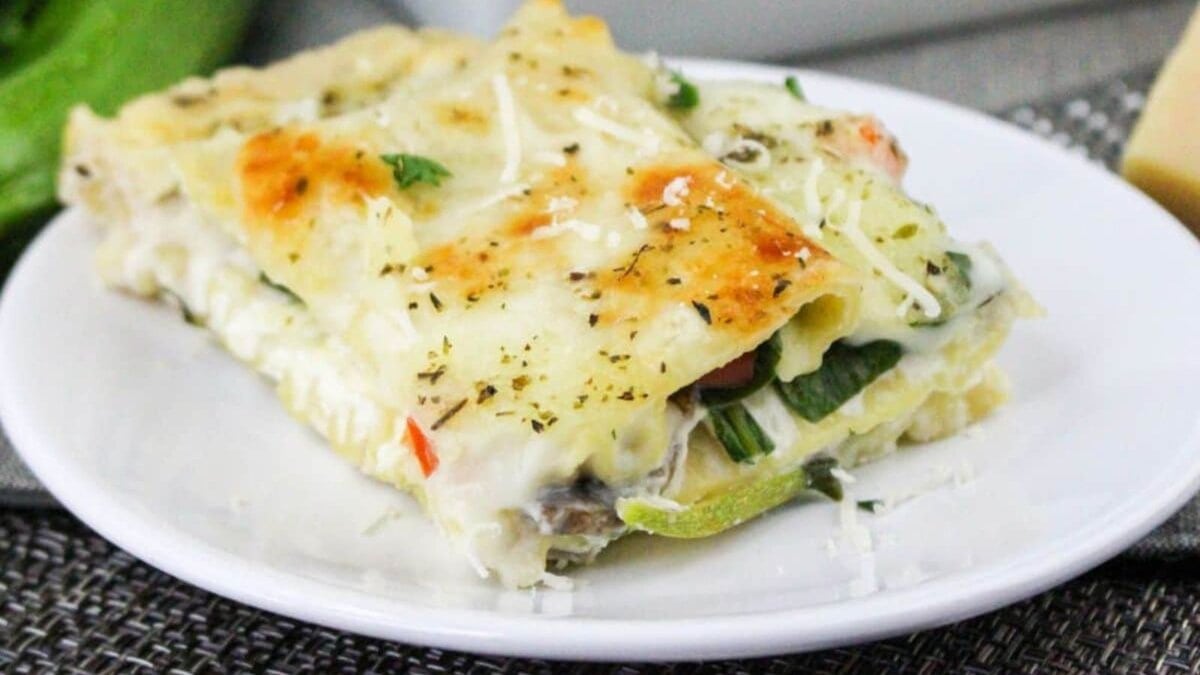 A slice of vegetable lasagna on a white plate, featuring layers of pasta, cheese, and vegetables, garnished with herbs and grated cheese.